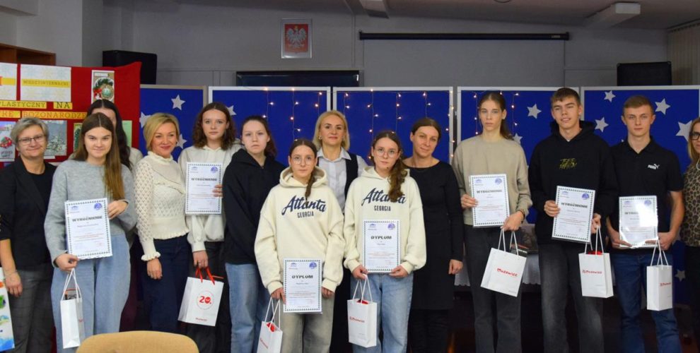 Na zdjęciu znajdują uczestnicy XIII Międzyinternackiego konkursu plastycznego na „Kartkę Bożonarodzeniową” wraz z opiekunami. W dłoniach trzymają dyplomy i nagrody. Z tyłu znajduje się czerwona tablica z kartkami konkursowymi i niebieska tablica z migającymi światełkami i białymi gwiazdkami.