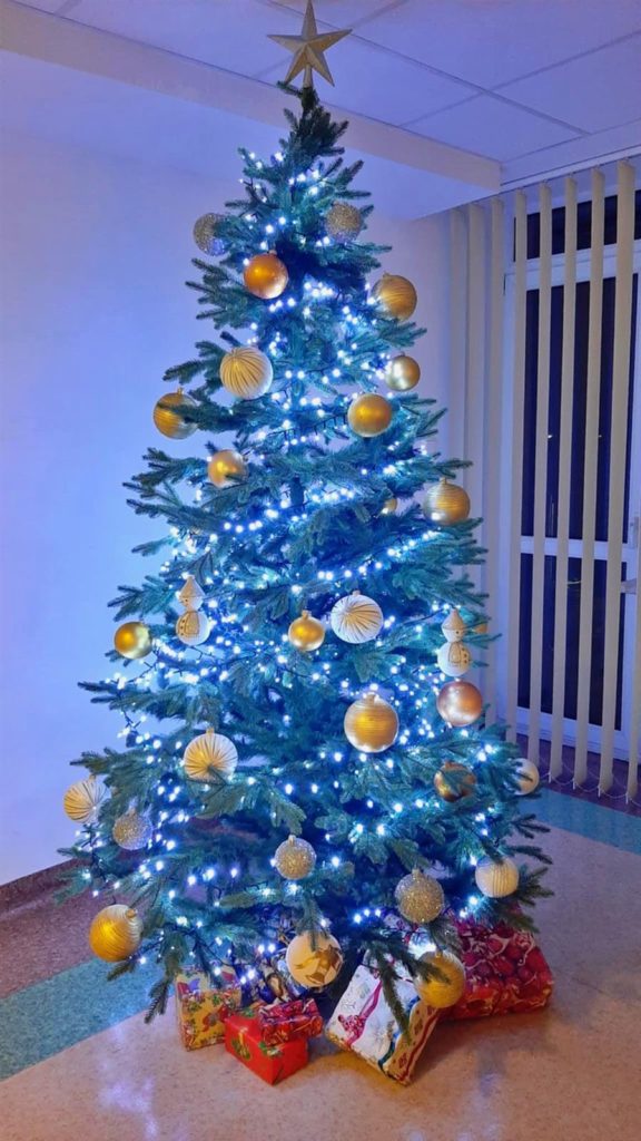 Na pierwszym planie znajduje się zielona choinka przystrojona kolorowymi bombkami, światełkami. Pod choinką znajdują się prezenty.