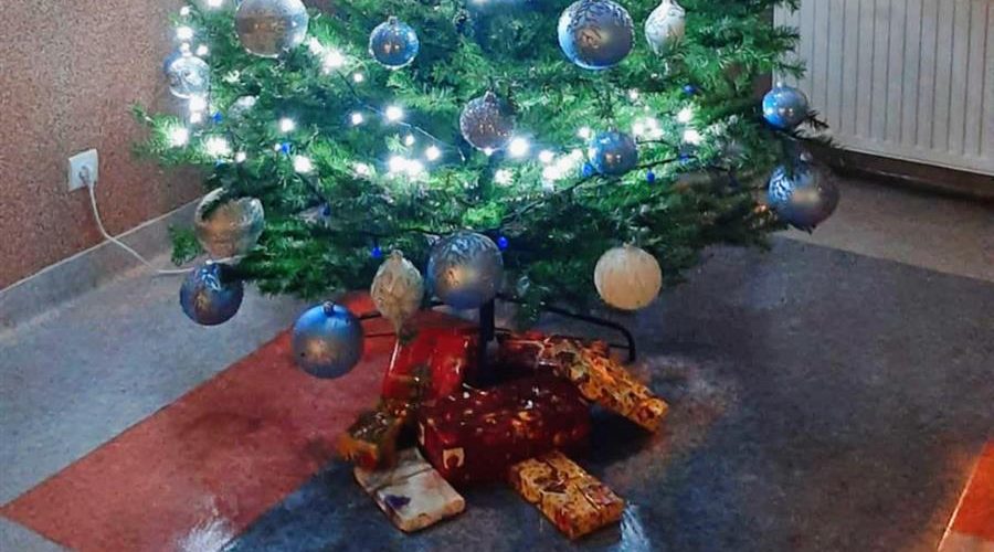 Na pierwszym planie znajduje się zielona choinka przystrojona kolorowymi bombkami, światełkami. Pod choinką znajdują się prezenty