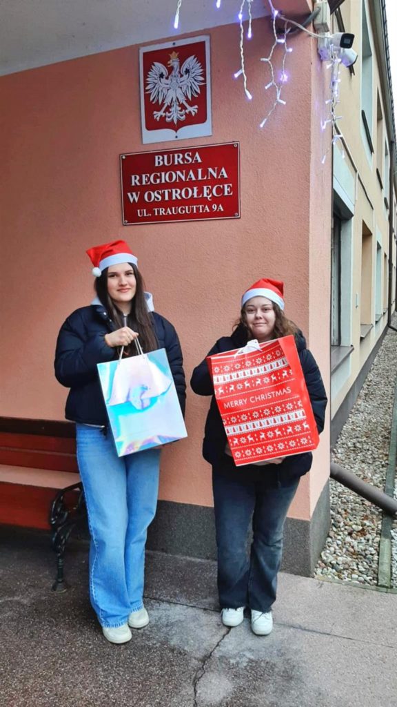 Dwie wychowanki bursy stoją przed placówką bursy. Na głowie mają czapki Mikołaja, w rękach trzymają torby świąteczne w których znajdują się dary zebrane podczas akcji pt. „I ty możesz zostać św. Mikołajem”.
