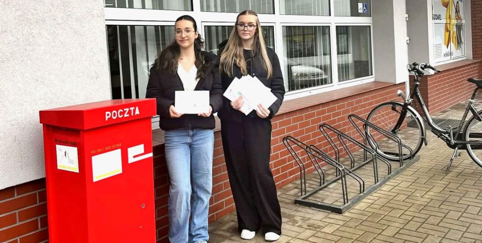 Dwie młode osoby stoją przed budynkiem Poczty Polskiej obok czerwonej skrzynki pocztowej. Trzymają w rękach świąteczne kartki i koperty, biorą udział w akcji „Idą Święta – nie o SMS-ie, lecz o kartce pamiętaj”