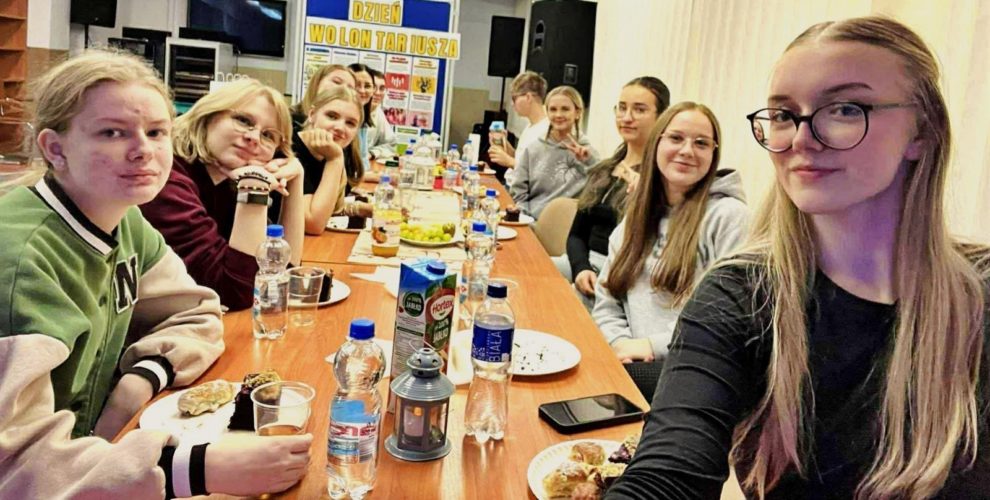 Młodzież siedząca przy długim stole w świetlicy bursy podczas spotkania z okazji Dnia Wolontariusza, wspólnie spędzająca czas przy przekąskach i napojach.