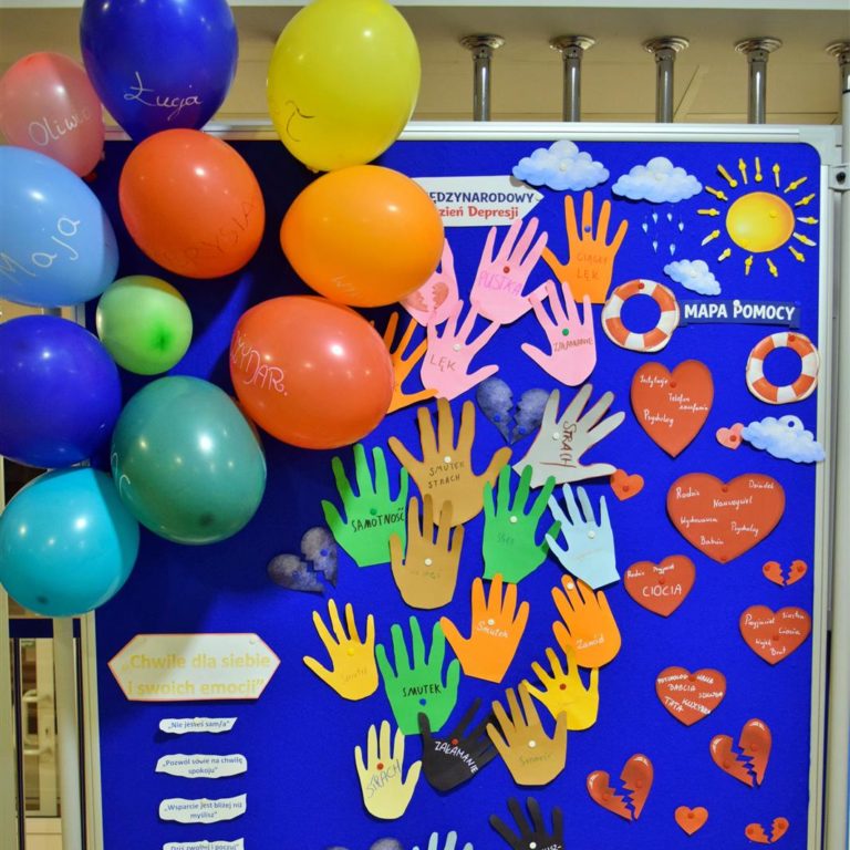 Na zdjęciu widać wystawę prac wychowanków, poświęconą depresji (napis: „Międzynarodowy Dzień Depresji”). Tablica jest ozdobiona kolorowymi wycięciami w kształcie dłoni z nazwami emocji (np. smutek, lęk, samotność), sercami oraz rysunkami chmur i słońca. Po lewej stronie znajdują się kolorowe balony z imionami.