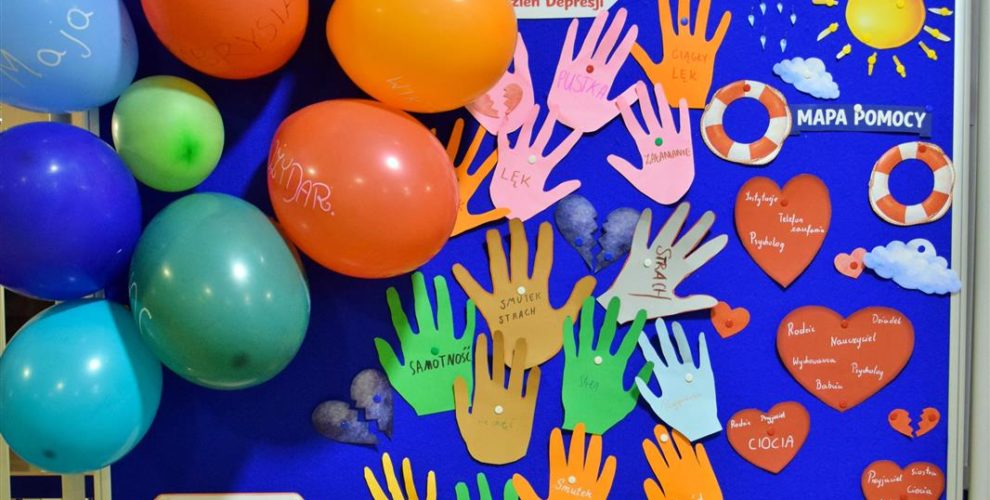 Na zdjęciu widać wystawę prac wychowanków, poświęconą depresji (napis: „Międzynarodowy Dzień Depresji”). Tablica jest ozdobiona kolorowymi wycięciami w kształcie dłoni z nazwami emocji (np. smutek, lęk, samotność), sercami oraz rysunkami chmur i słońca. Po lewej stronie znajdują się kolorowe balony z imionami.