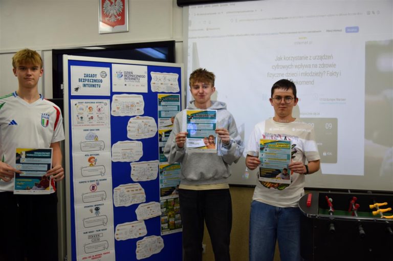 Trzech wychowanków bursy stoi przy tablicy, na której znajduje się wystawa z okazji obchodów Dnia Bezpiecznego Internetu 2026 r. W rękach trzymają plakaty edukacyjne: nt. „Weź nie hejtuj – to przemoc. Widzisz hejt? Co robić? Jak pomóc? oraz Dotyka Cię hejt? Trudno Ci o tym mówić? Na ścianie na rzutniku wyświetlany jest webinar pt. „Jak korzystanie z urządzeń cyfrowych wpływa na zdrowie fizyczne młodzieży?