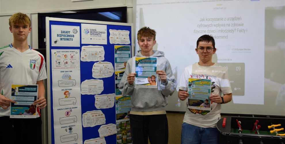Trzech wychowanków bursy stoi przy tablicy, na której znajduje się wystawa z okazji obchodów Dnia Bezpiecznego Internetu 2026 r. W rękach trzymają plakaty edukacyjne: nt. „Weź nie hejtuj – to przemoc. Widzisz hejt? Co robić? Jak pomóc? oraz Dotyka Cię hejt? Trudno Ci o tym mówić? Na ścianie na rzutniku wyświetlany jest webinar pt. „Jak korzystanie z urządzeń cyfrowych wpływa na zdrowie fizyczne młodzieży?