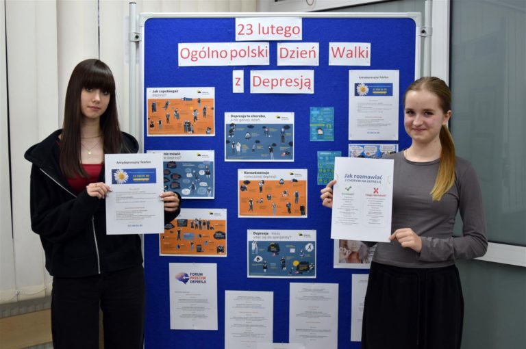 Dwie wychowanki bursy stoją przy niebieskiej tablicy na której znajduje się wystawa z okazji Ogólnopolskiego Dnia Walki z Depresję. W rękach trzymają materiały edukacyjne nt. Antydepresyjny Telefon oraz Jak rozmawiać z chorym na depresję?.