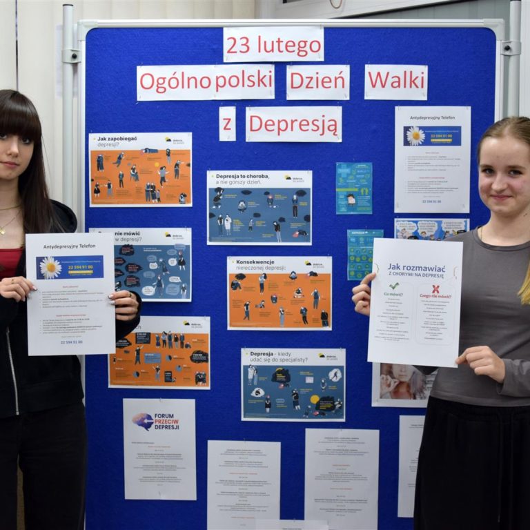 Dwie wychowanki bursy stoją przy niebieskiej tablicy na której znajduje się wystawa z okazji Ogólnopolskiego Dnia Walki z Depresję. W rękach trzymają materiały edukacyjne nt. Antydepresyjny Telefon oraz Jak rozmawiać z chorym na depresję?.