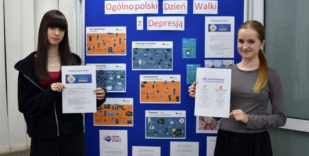 Dwie wychowanki bursy stoją przy niebieskiej tablicy na której znajduje się wystawa z okazji Ogólnopolskiego Dnia Walki z Depresję. W rękach trzymają materiały edukacyjne nt. Antydepresyjny Telefon oraz Jak rozmawiać z chorym na depresję?.