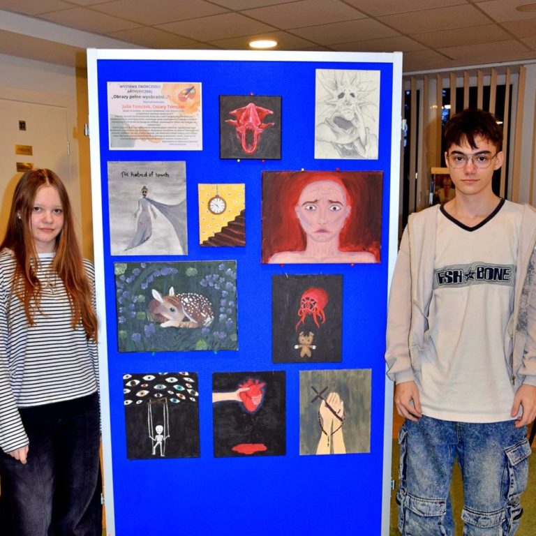 Dwoje wychowanków bursy stoi przy niebieskiej tablicy. Na tablicy znajduje się wystawa prac wychowanków (obrazów), na której widnieją elementy ludzkie (twarze, dłonie), zwierzęta, schody, zegar ścienny.