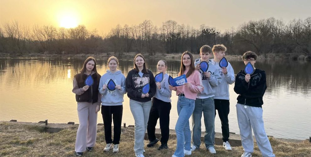 Grupa młodych osób pozuje do zdjęcia na tle wody i zachodu słońca. W rękach trzymają materiały promocyjne z akcji: Światowy Dzień Wody.