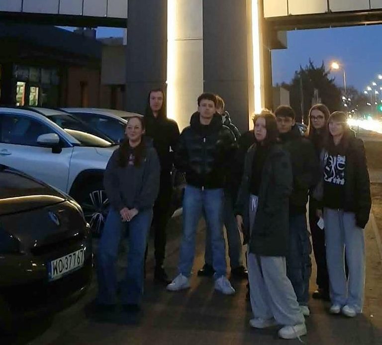 Dziesięcioro wychowanków stoi przed wejściem do Muzeum Żołnierzy Wyklętych w Ostrołęce. Za nimi znajduje się oświetlona kładka piesza, ulica oświetlona latarniami. Po lewej stronie stoją zaparkowane samochody. Na niebie zapada wieczór.