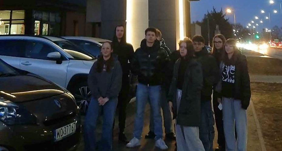 Dziesięcioro wychowanków stoi przed wejściem do Muzeum Żołnierzy Wyklętych w Ostrołęce. Za nimi znajduje się oświetlona kładka piesza, ulica oświetlona latarniami. Po lewej stronie stoją zaparkowane samochody. Na niebie zapada wieczór.