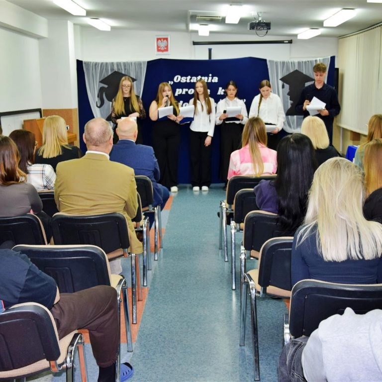 Grupa młodzieży stoi na scenie podczas uroczystego pożegnania maturzystów, a przed nimi siedzi publiczność.