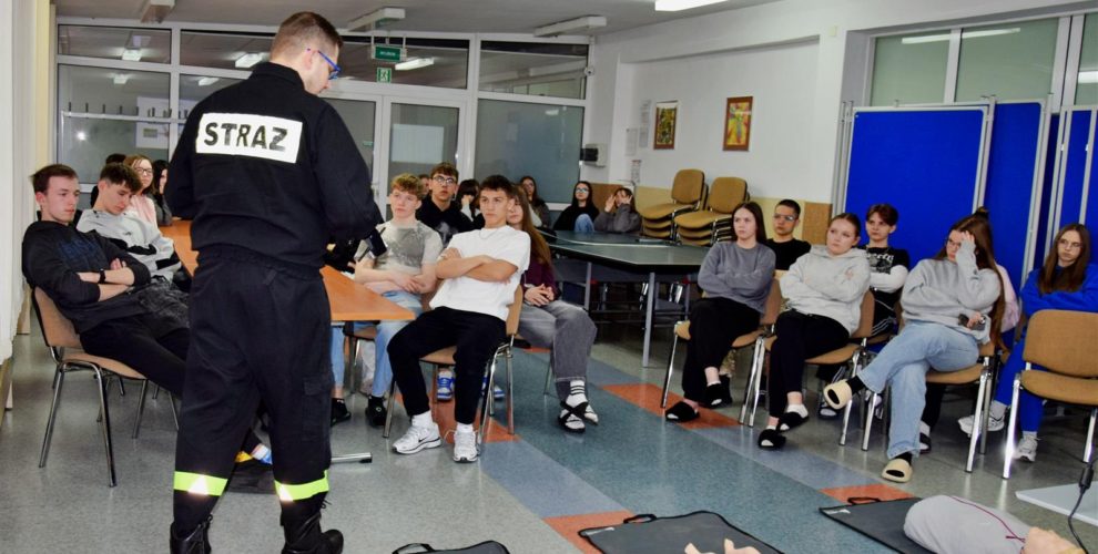 Wychowankowie siedzący na krzesłach oraz słuchający Strażaka, na podłodze leżące fantomy