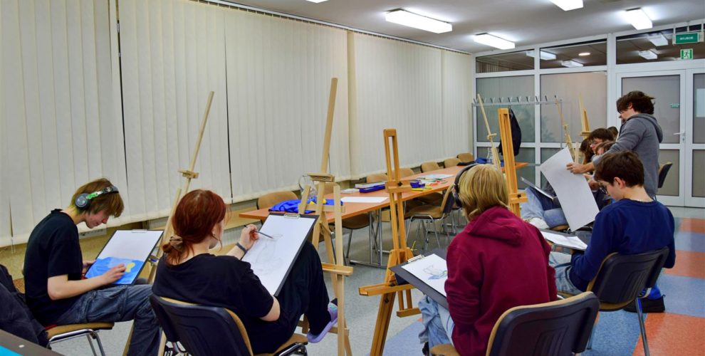 Dziewięcioro wychowanków siedzi przy sztalugach malarskich. Na kartkach rysują ołówkiem i pastelami martwą naturę (dzbanek który stoi na materiale, przy nim leży przekrojona na pół cytryna). W oddali widać krzesła i stoły na których stoją dwie ekspozycje: dzbanek na niebieskim i granatowym materiale, z boku leży przekrojona cytryna. Przy wychowankach stoi Pan prowadzący warsztaty pokazujący i tłumaczący wychowankom sposoby malowania.