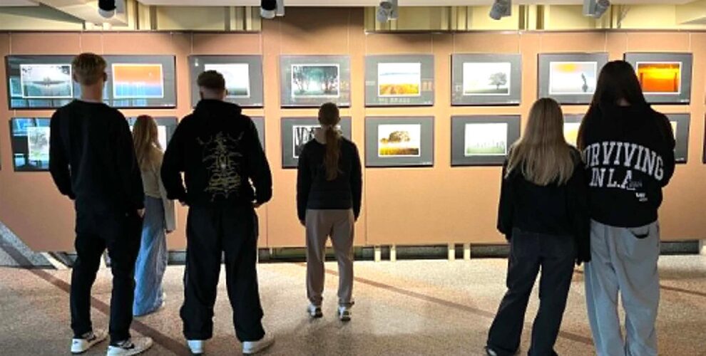 Grupa młodzieży ogląda wystawę fotografii na korytarzu wystawowym. Osoby stoją tyłem do obiektywu, skupiając uwagę na prezentowanych pracach, które przedstawiają krajobrazy. Oświetlenie podkreśla szczegóły eksponatów, tworząc klimatyczną atmosferę.