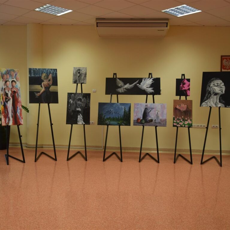 Widok na całą ekspozycję kilkunastu prac malarskich ustawionych rzędem na sztalugach wzdłuż ściany czytelni Biblioteki Pedagogicznej w Ostrołęce