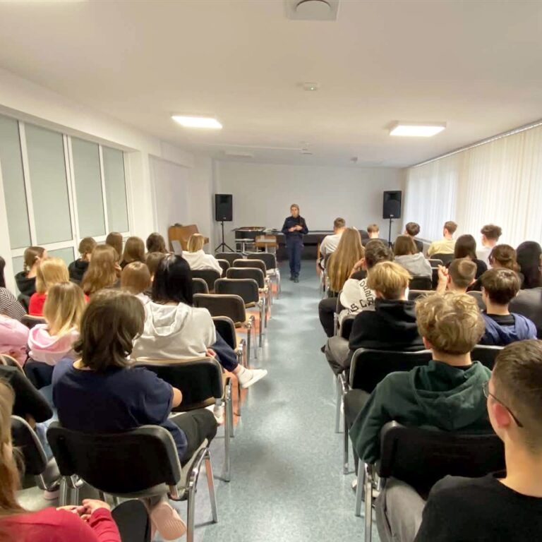 Na pierwszym planie znajdują się wychowankowie bursy. Siedzą na krzesłach, skierowani tyłem do wejścia, przodem do funkcjonariusza Komendy Miejskiej Policji w Ostrołęce. Zaproszony gość przeprowadza z wychowankami bursy pogadankę profilaktyczną. Spotkanie odbywa się w świetlicy Bursy Regionalnej w Ostrołęce.